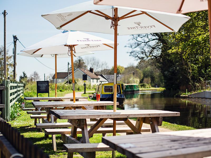 Picnic benches under parasols on the canal bank outside the pub, narrowboats moored behind